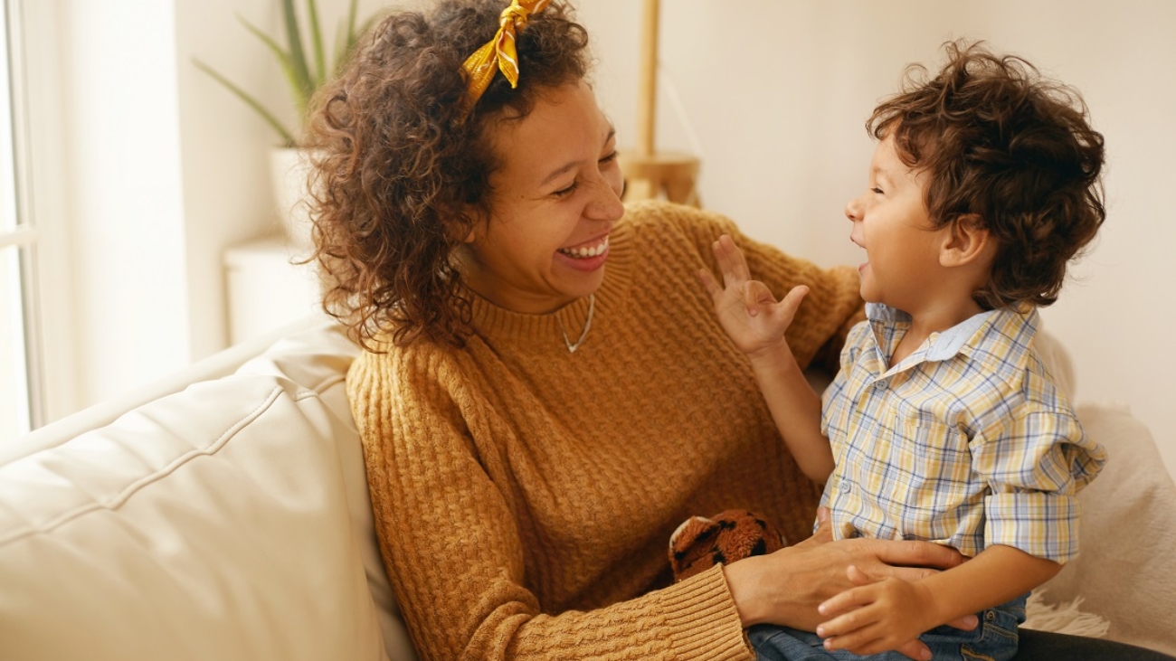 Indoor shot of happy young Hispanic woman with brown wavy hair relaxing at home embracing her adorable toddler son. Cheerful mother bonding with infant son, sitting on sofa in living room, laughing