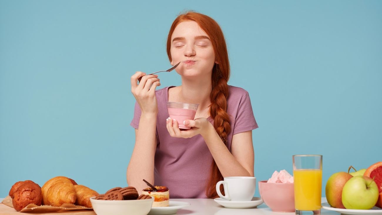 Nice red-haired girl trying tasting cherry yogurt wirh a teaspoon, closed her eyes from pleasure, sitting at the table during lunch, pastries on the table and fresh fruit, against a blue background.