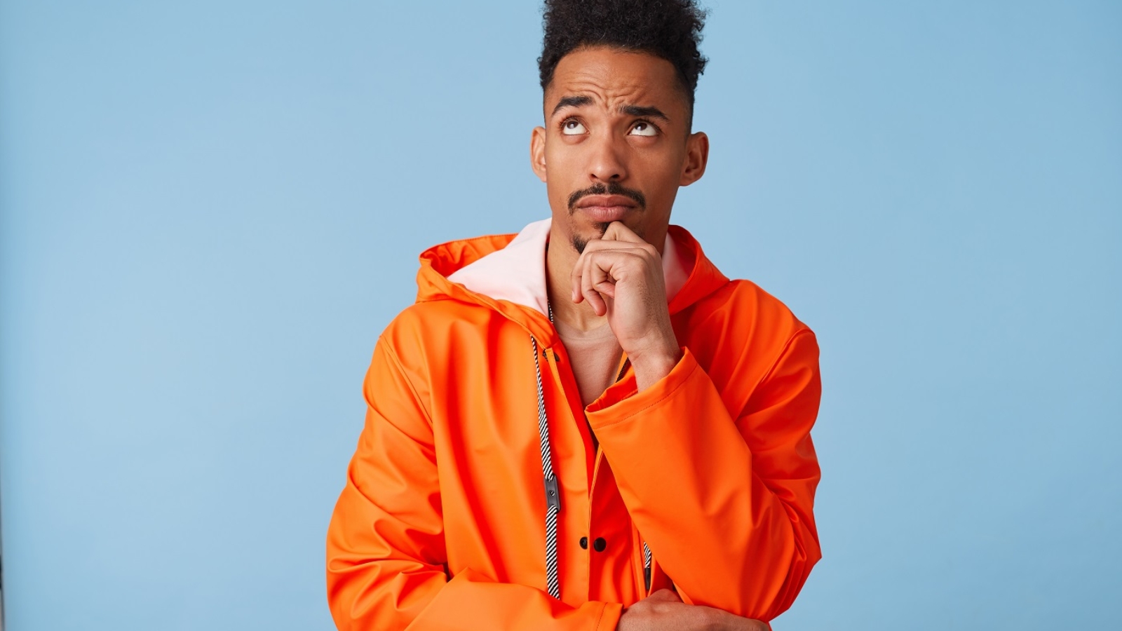 Portrait of young african american dark skinned man in orange rain coat, holds his chin, looks up and thinks about plans for the future stands over blue background.