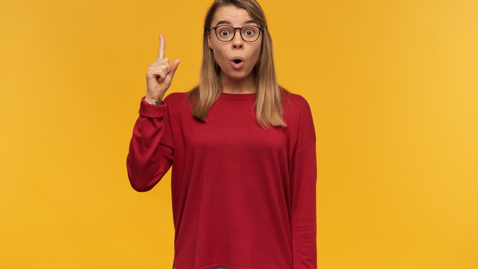 Young girl looking inspired, with rounded mouth, has blond hair. Wearing glasses. Pointing up with index finger on left hand like having a great idea. Looking camera isolated over yellow background