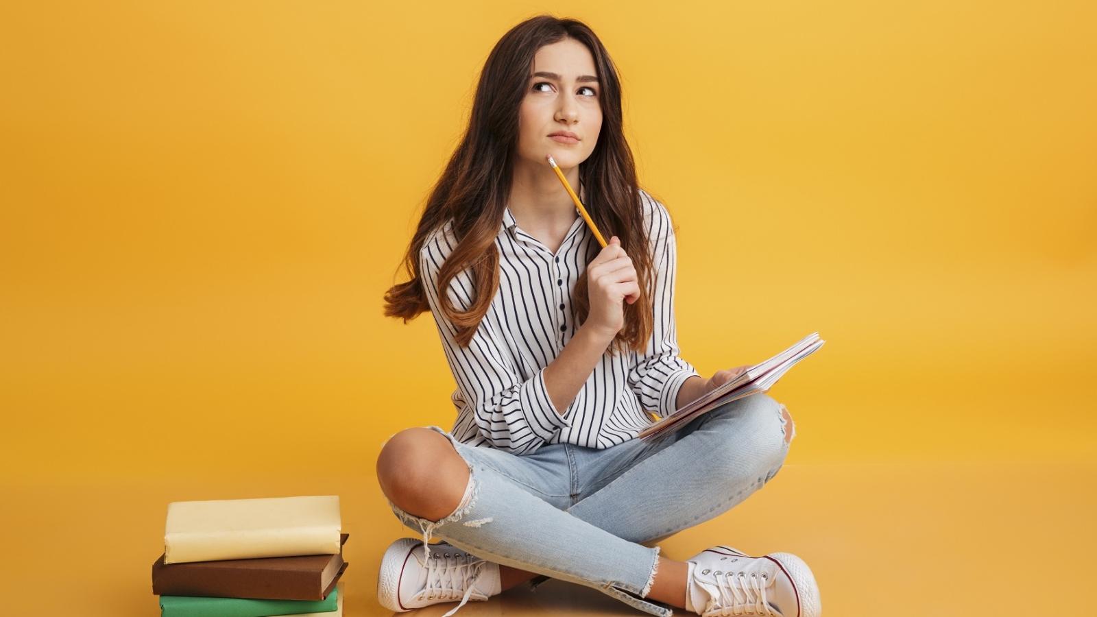 Portrait of a pensive young girl making notes while sitting with books isolated over yellow background