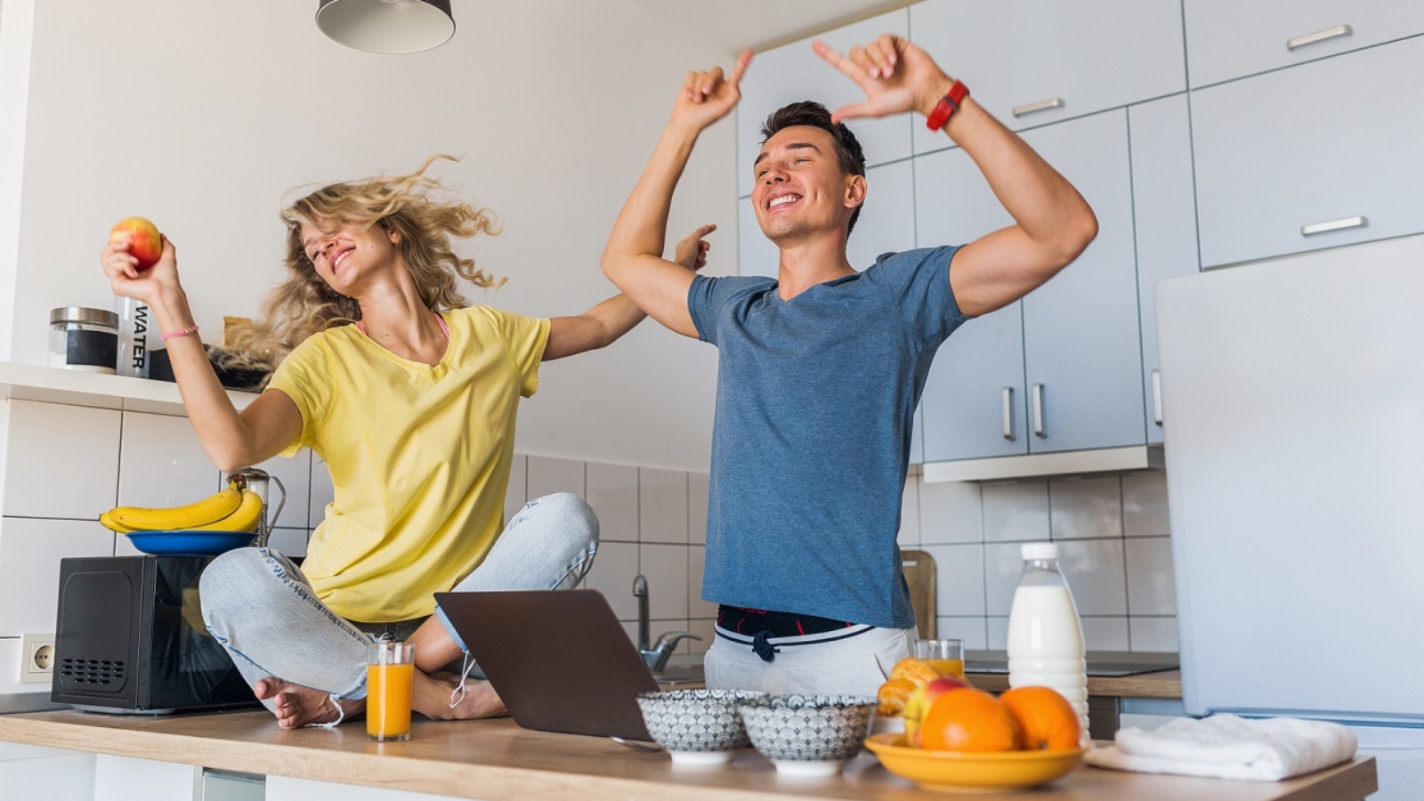 young attractive couple of man and woman cooking breakfast together in morning at kitchen, happy family, romantic, healthy food, freeelancer at work on laptop, dancing to music, having fun, laughing